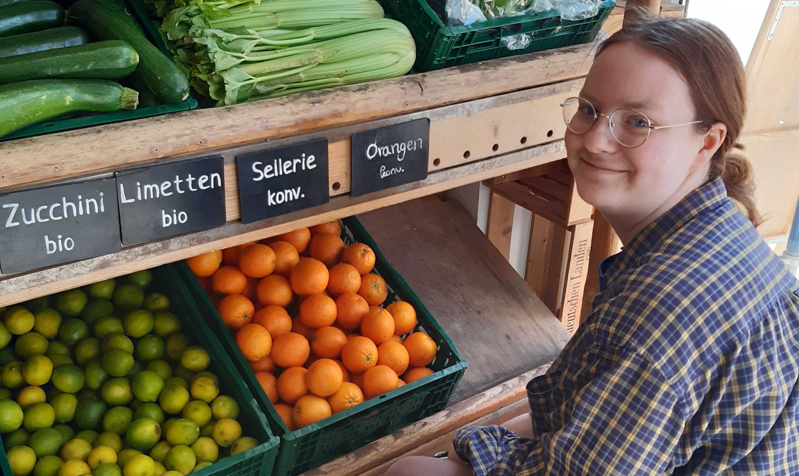 Foto von Alina, die vor prall mit Gemüse gefüllten Regalen im Laden sitzt.