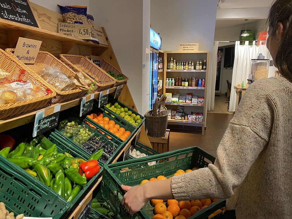 Zu sehen ist in einem Ladengeschäft eine junge Frau im Halprofil, die eine Kiste mit Gemüse hält. Links von ihr ein prall mit unterschiedlichem Gemüse und Obst gefülltes Regal.